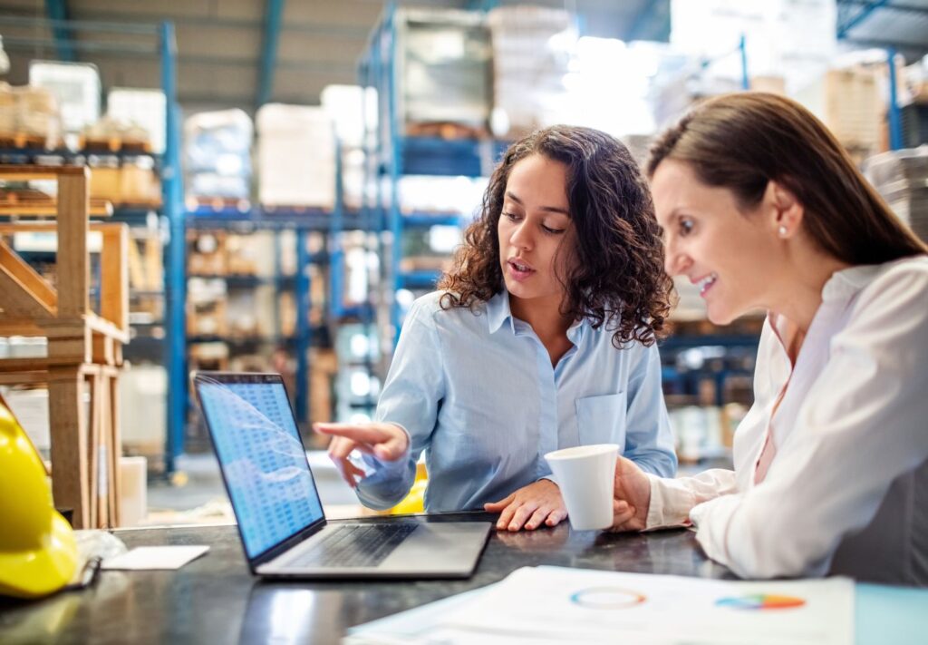 two women sitting at desk in warehouse looking at a b-stock inventory on  laptop